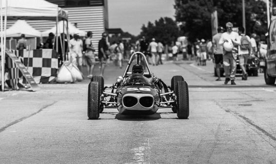Photo en noir et blanc d'une ancienne Formule Junior sur les paddocks de Dijon-Prenois, capturant l'essence classique de la voiture de course vintage dans un environnement de course légendaire
