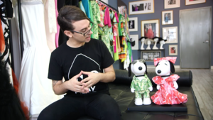 a man sits next to two dog figures, one ina green suit jacket and one in a pink dress