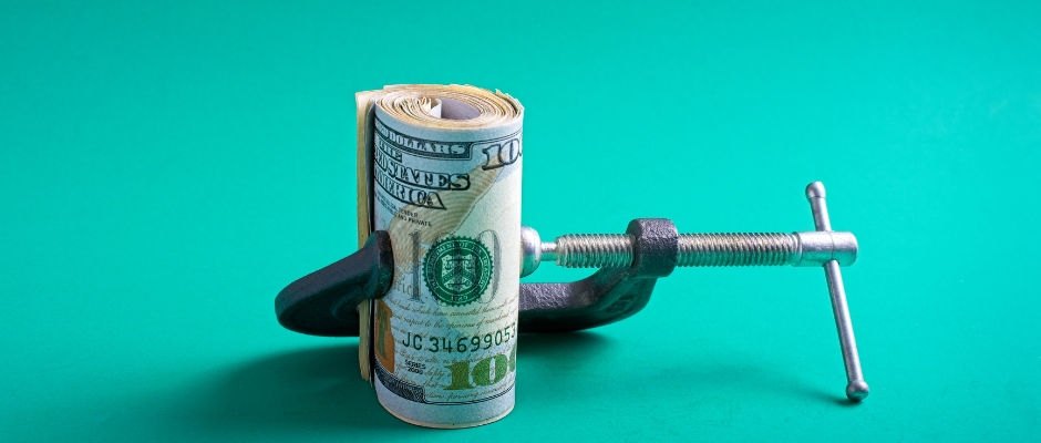 Rolled U.S. dollar bills being tightened in a metal clamp, symbolizing financial pressure, rising costs, and constrained budgets.