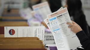 A person reviews and sorts official election ballots at a table, with other workers and ballot envelopes visible in the background.