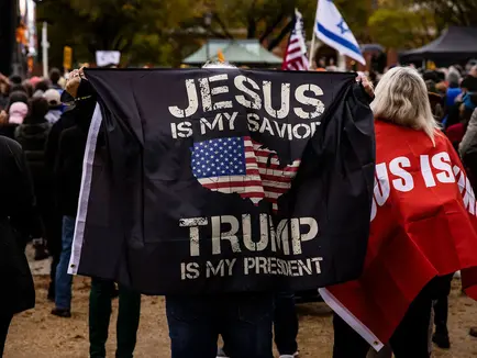 Man wearing a Christian nationalist flag draped over his shoulders at an outdoor gathering