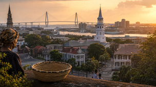 A Black woman in a patterned headwrap overlooks Charleston at golden hour beside a traditional sweetgrass basket, with historic homes, church steeples, the harbor and Ravenel Bridge in the distance, symbolizing the city’s enduring Gullah Geechee and African American heritage.