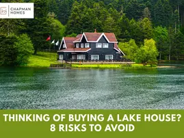 Chapman Homes blog featured image showing a large dark lakefront home surrounded by evergreen trees reflected in calm lake water, titled "Will You Regret Buying a Lake House? 8 Risks to Avoid"