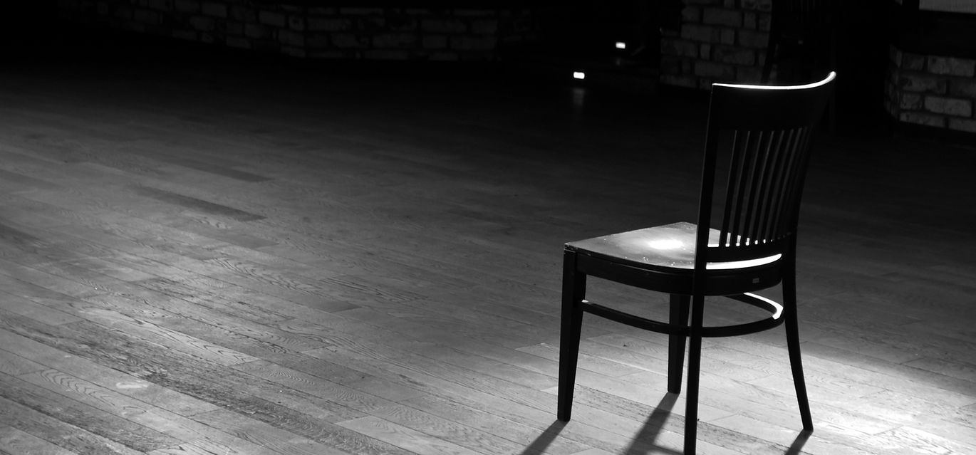 A wooden chair standing in the middle of an empty stage under the light of soffits, in bla