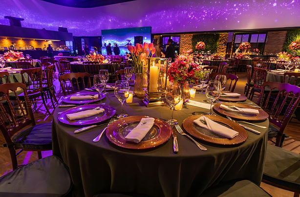 An elegant gala-style event with round tables set with gold chargers, folded white napkins, wine glasses, and floral centerpieces. The room is softly lit with purple ambient lighting, creating a sophisticated, celebratory atmosphere.