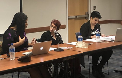 Liat sits at a long table as a part of a panel discussion with two other people. Liat, a white presenting woman with short red hair and glasses, is looking at a Black woman that appears to be asking her a question or discussing a point with her.