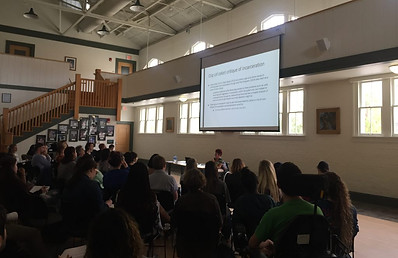 Image of Liat Ben-Moshe sitting in her wheelchair addressing a crowd of people at the University of Florida, Gainesville. She is presenting a critique of incarceration on a powerpoint slide.