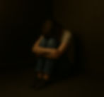 Person sitting on the floor in a dark room, hugging knees. Wearing a gray shirt and jeans. Mood is somber and introspective.