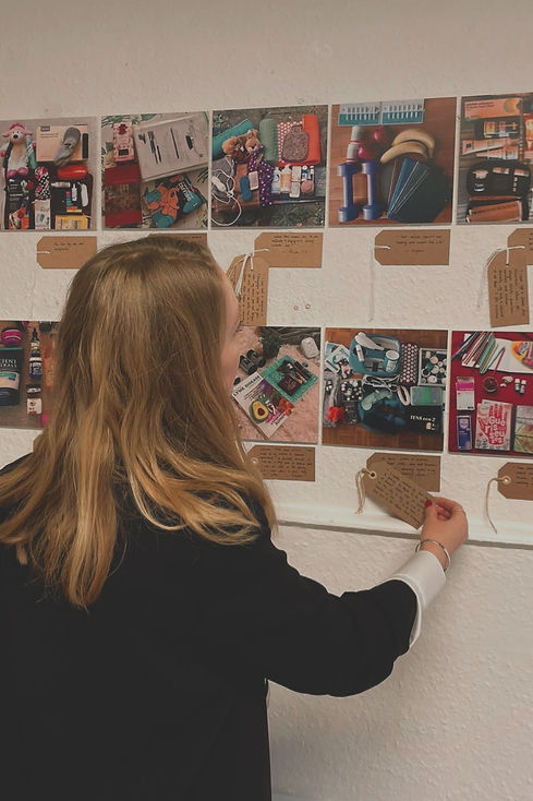 A photograph of Angela in a white shirt and black blazer and long blonde hair reaching up to a board of flatlay images of items that assist in the day-to-day lives of those with chronic illness. Underneith each image is a brown packaging label with handwritten text to explain the contents of the photograph.