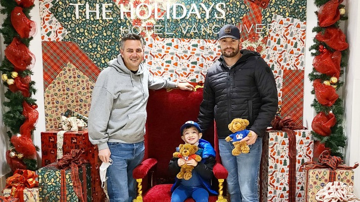 Two men stand in front of Christmas display with young boy sitting in the middle, holding a teddy bear | Community Key