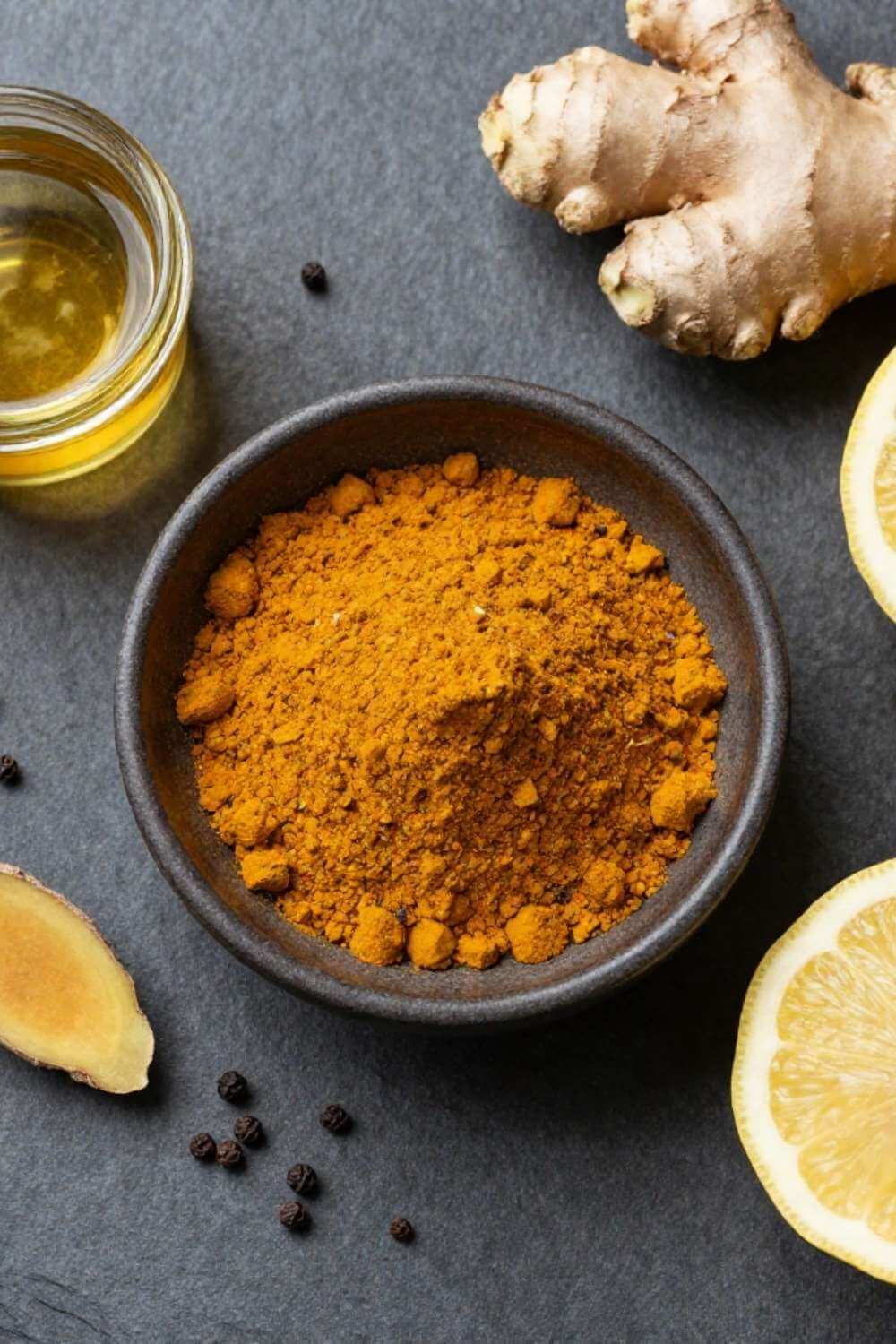 Ground turmeric powder in a small bowl with a scattering of black peppercorns on a stone surface