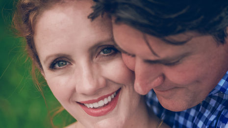 a woman wearing a heart shaped necklace smiles while being hugged by a man