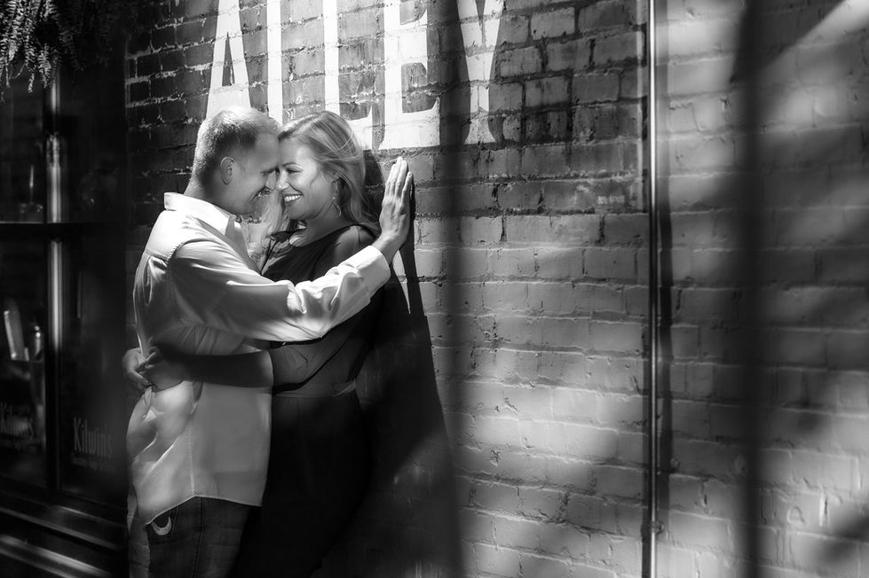 a black and white photo of a man and woman hugging in front of a brick wall with the word alley painted on it