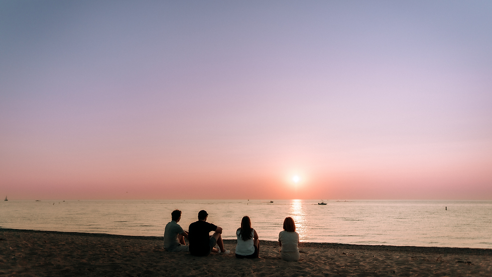 Four people sit on a sandy beach watching a pink sunset over calm water.