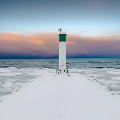 Lighthouse at the end of a pier. Snow covered ground and icy water with a pink and purple sky.