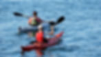 Two kayakers paddle on calm blue water, one in a red kayak and one in a white kayak, both wearing life vests.