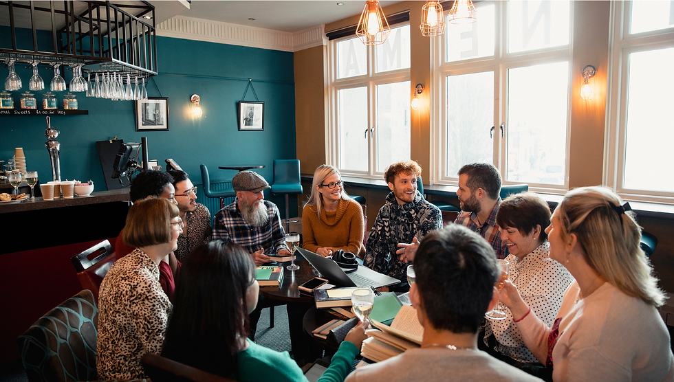 Group of book club members chatting around a café table with laptop, books and drinks in a bright teal-walled room with large windows.