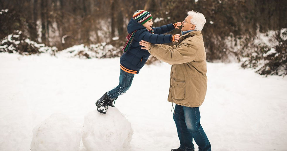 A child balances on snowballs, reaching out to an older man in a snowy forest. Both wear winter coats, the scene is cheerful and playful.