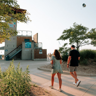 A man and woman walk on a sunny path near a modern building with trees and a balloon in the sky, creating a relaxed, summery vibe.