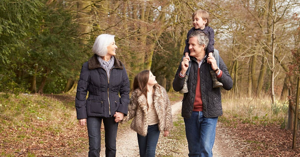 A family enjoying a walk in a forest. An elderly woman holds a girl's hand, while a man carries a boy on his shoulders. All are smiling.