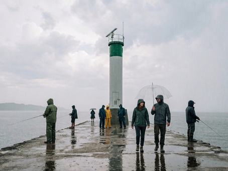 Rainy pier with several people in raincoats fishing and walking beside a green-and-white lighthouse under gray skies.