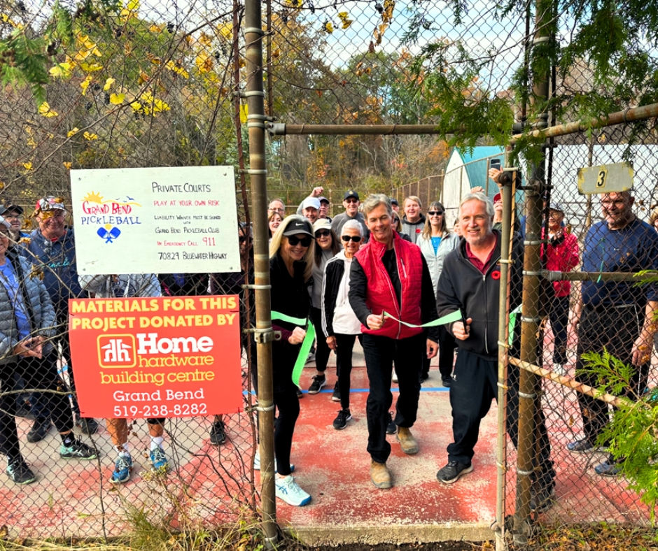 People at a ribbon-cutting ceremony on a sunny day at a pickleball court surrounded by trees. Signs for Grand Bend Pickleball and Home Hardware.