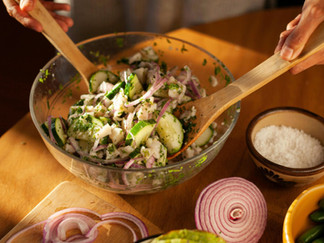 Hands mix a salad with cucumber, onion, and herbs in a glass bowl. Nearby are sliced onions, salt, garlic, and chilies on a wooden table.