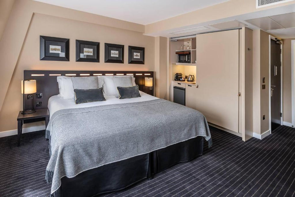 Chambre d’hôtel moderne avec grand lit gris, lampes de chevet et kitchenette intégrée, ambiance calme.