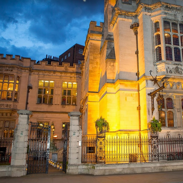 Façade d’un bâtiment historique éclairé en jaune au crépuscule, derrière une grille noire, sous un ciel bleu orageux.