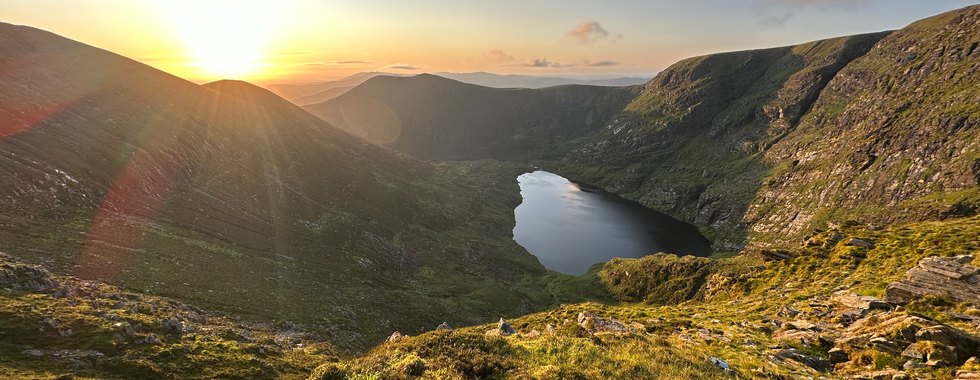 Im Vordergrund ist die Vulkanähnliche Form des Mangerton zu sehen, mit dem See in der Mitte. Der Ausblick zeigt Silhouetten weiterer Bergketten hinter denen gerade die Sonne aufgegangen ist, sie scheint hell gelb-orange am Himmel.