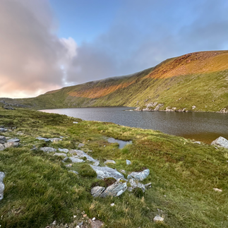 Ein Bergsee liegt ruhig zwischen den grünen Hügeln. Die Sonne bescheint die gegenüberliegende Uferseite