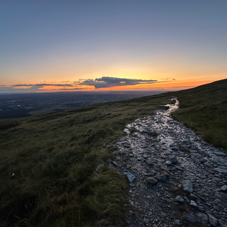 Ein Weg schlängelt sich den Berg entlang, er glitzert vor nässe. Hinter dem Berg leuchtet der Himmel orange.