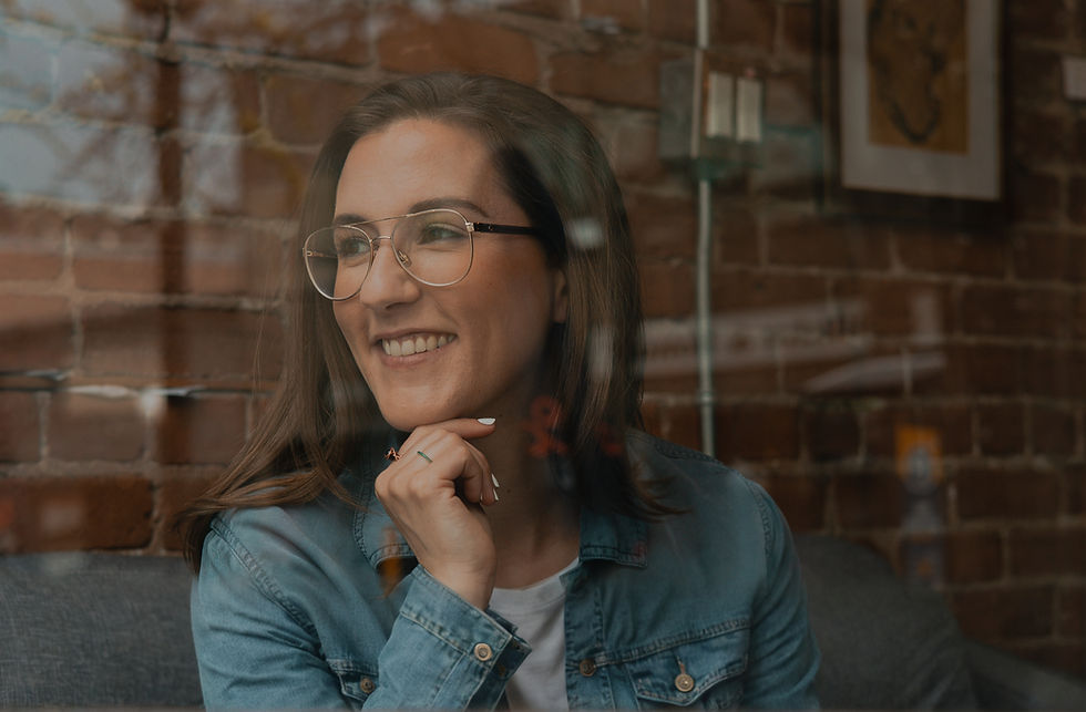 A smiling woman with brown hair and gold glasses sitting on a grey couch with a brick wall behind her. She is looking away from the camera, her hand on her chin.