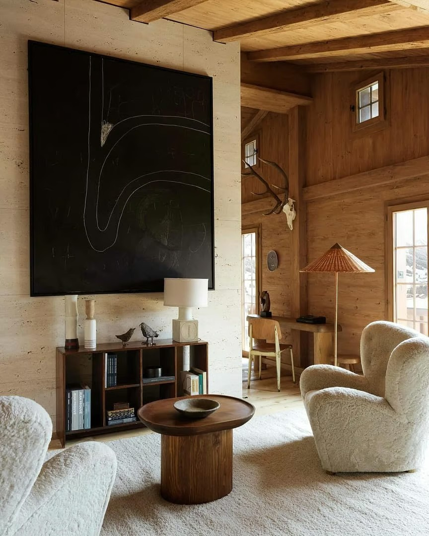 A modern cabin living room featuring a blend of rustic and contemporary elements. The focal point is a large, dramatic black artwork mounted on a wooden accent wall. Natural light streams through expansive windows, illuminating a cozy seating area with curvy furniture.