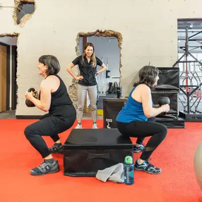 Two women performing goblet squats with kettlebells on red turf at Zenith Performance and Wellness, coached by a smiling trainer standing behind them near a soft plyo box.