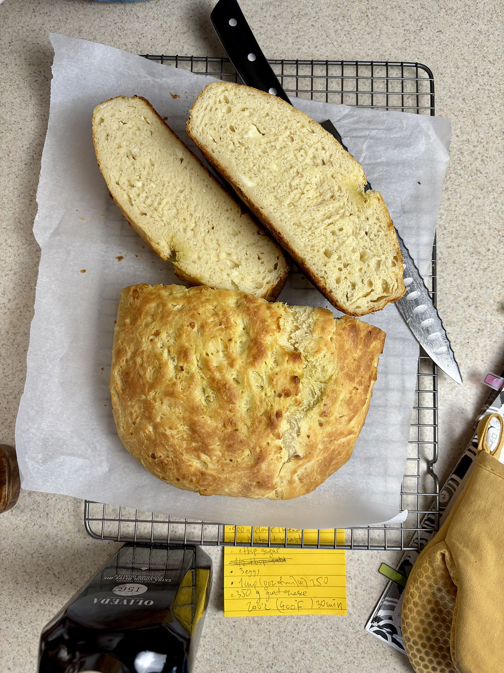 Loaf of bread on cooling rack, two slices cut, knife nearby. Yellow recipe card and bottle in background. Soft lighting, neutral tones.