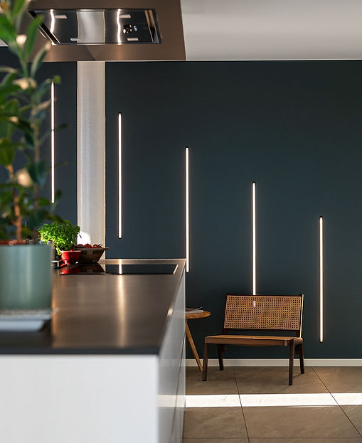 Ein modernes Wohnraumdesign mit einer dunkelblauen Wand, in die vertikale LED-Lichtleisten integriert sind. Diese sorgen für stilvolle Akzente und schaffen eine warme, einladende Atmosphäre. Im Vordergrund befindet sich eine moderne Küche mit Dekorationen und Pflanzen. Hörl Lichttechnik ist für die präzise Umsetzung der Lichtakzente verantwortlich, die das elegante und zeitgenössische Design unterstreichen.