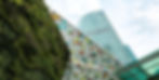 Green vertical garden wall and a colorful geometric glass canopy in front of a modern skyscraper against a cloudy sky.