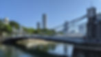 Suspension Cavenagh bdidge over calm river with city skyline in background. Clear blue sky, lush greenery. Two people on bridge's right side.