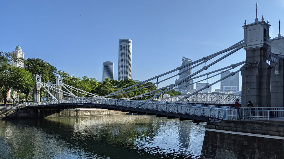 Suspension Cavenagh bdidge over calm river with city skyline in background. Clear blue sky, lush greenery. Two people on bridge's right side.