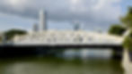 White arched cavenagh bridge over a river with tall skyscrapers in the background. Green trees on both sides under a partly cloudy sky.