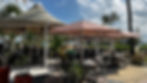 Outdoor cafe with striped umbrellas and potted plants. Patrons sit at tables under a sunny sky, with palm trees and a beach in the background.