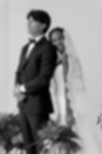 Bride in lace veil smiles, hugging groom in suit. He looks away. Black and white photo with floral decorations, capturing joy.