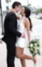A couple embraces lovingly outdoors; she wears a short white dress and holds a bouquet. The setting is urban with blurred traffic lights.