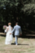 A bride and groom walk hand in hand through a sunlit park. The bride carries a bouquet, and the groom wears a light blue suit.