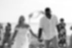 Bride and groom holding hands and laughing on a beach. Guests are clapping and taking photos. Sunny day with floral arch in background.