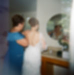 Bride in lace dress adjusts dress with help, reflected in mirror. Room with beige walls, flowers on counter. Calm, intimate mood.