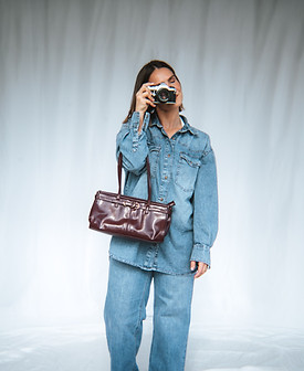 women in a blue outfit with a red purse holds a camera in front of her face with a white background
