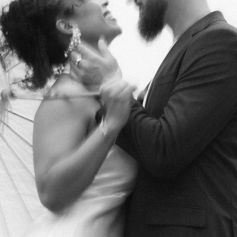 Couple in wedding attire embraces near a lakeside, with a white umbrella. The scene is joyful and romantic with a soft, misty backdrop.