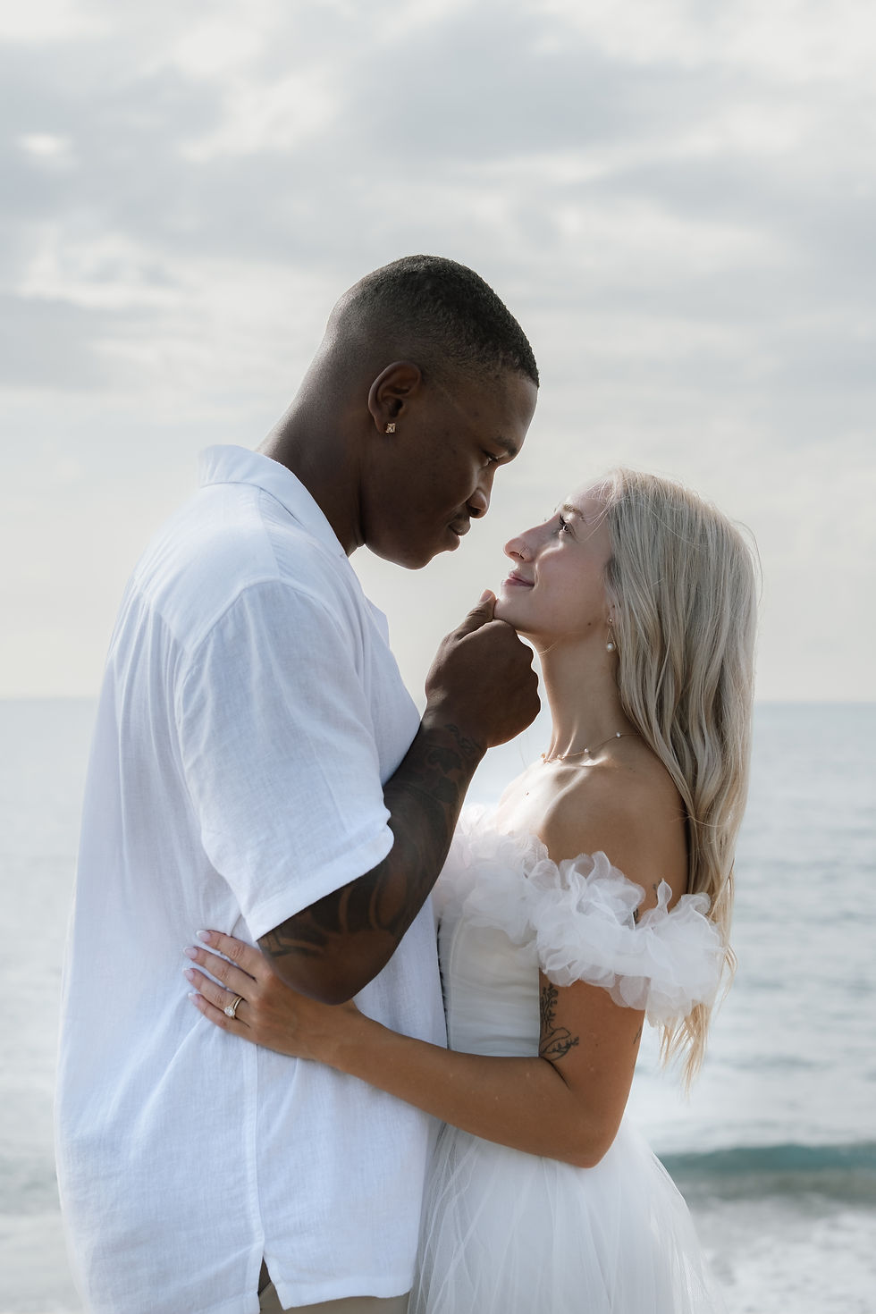 A couple stands closely on a beach, gazing lovingly at each other. The man wears a white shirt; the woman a white dress. Ocean in the background.